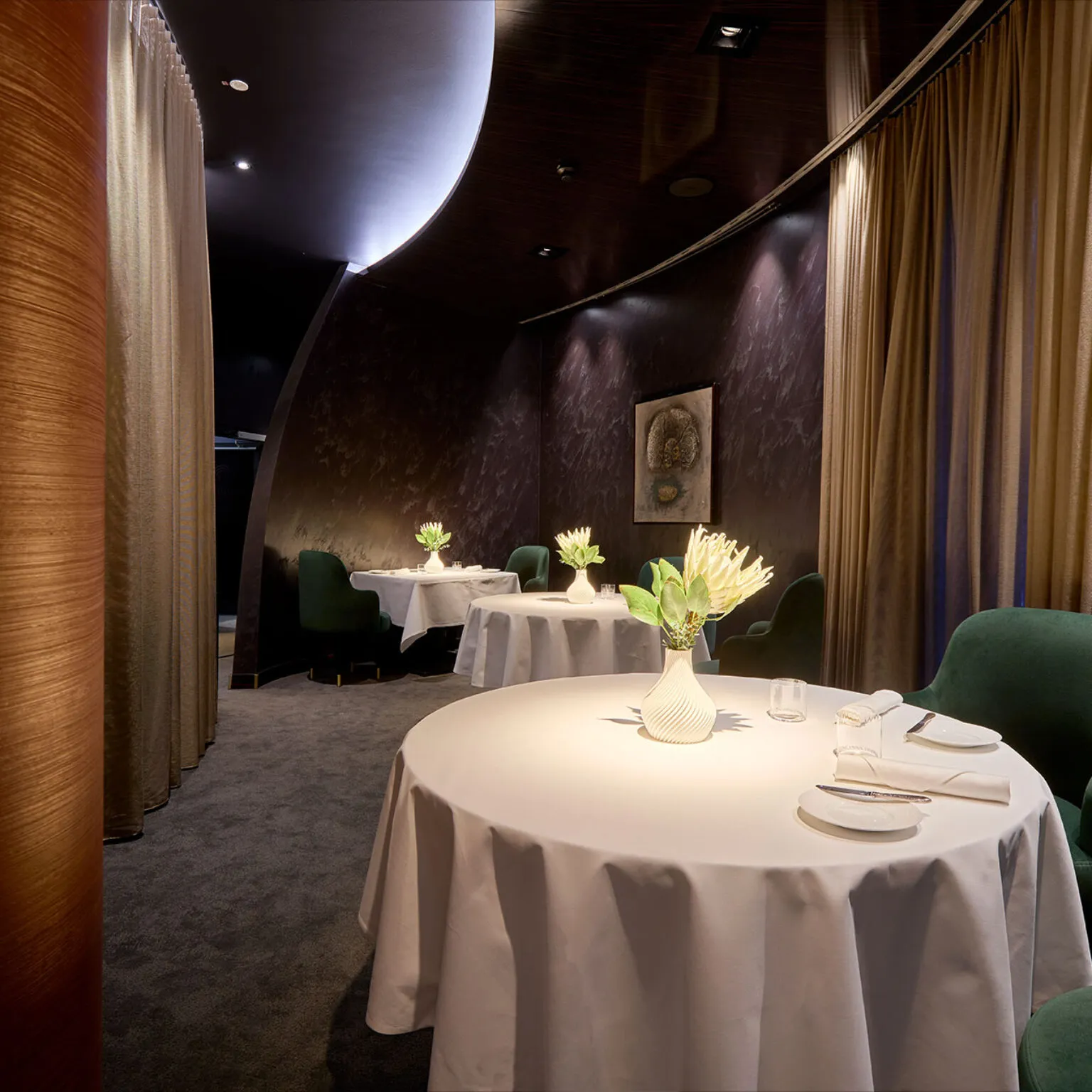 Innenansicht des stilvoll eingerichteten 2-Sterne-Restaurants Ammolite mit modernem Interieur in Grün- und Bronzetönen. Im Vordergrund stehen drei weiß gedeckte Tische mit Blumenvasen.