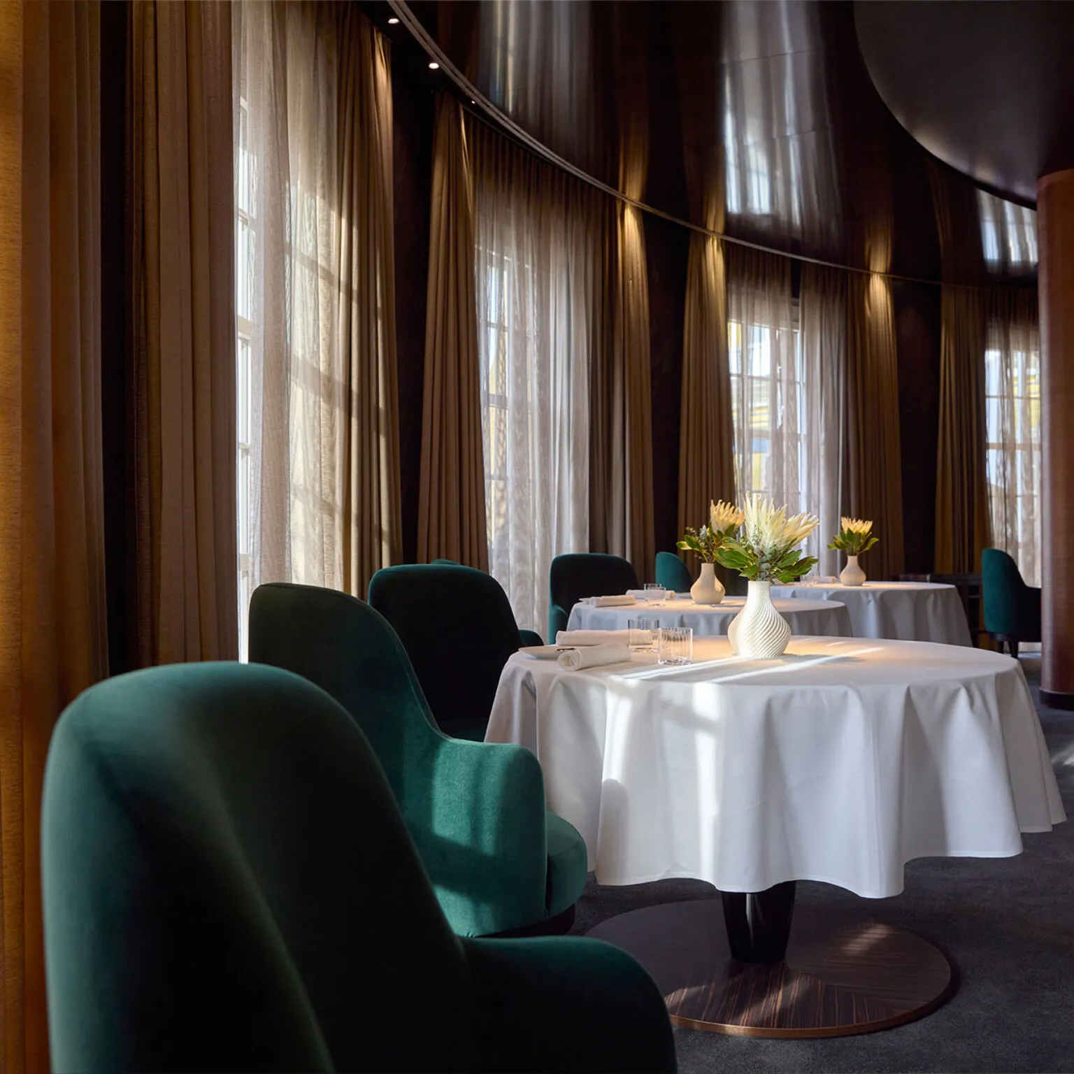 Innenansicht des Restaurants Ammolite – House of Light. Im Raum stehen weiß gedeckte Tische mit Blumenvasen und grüne Sessel.