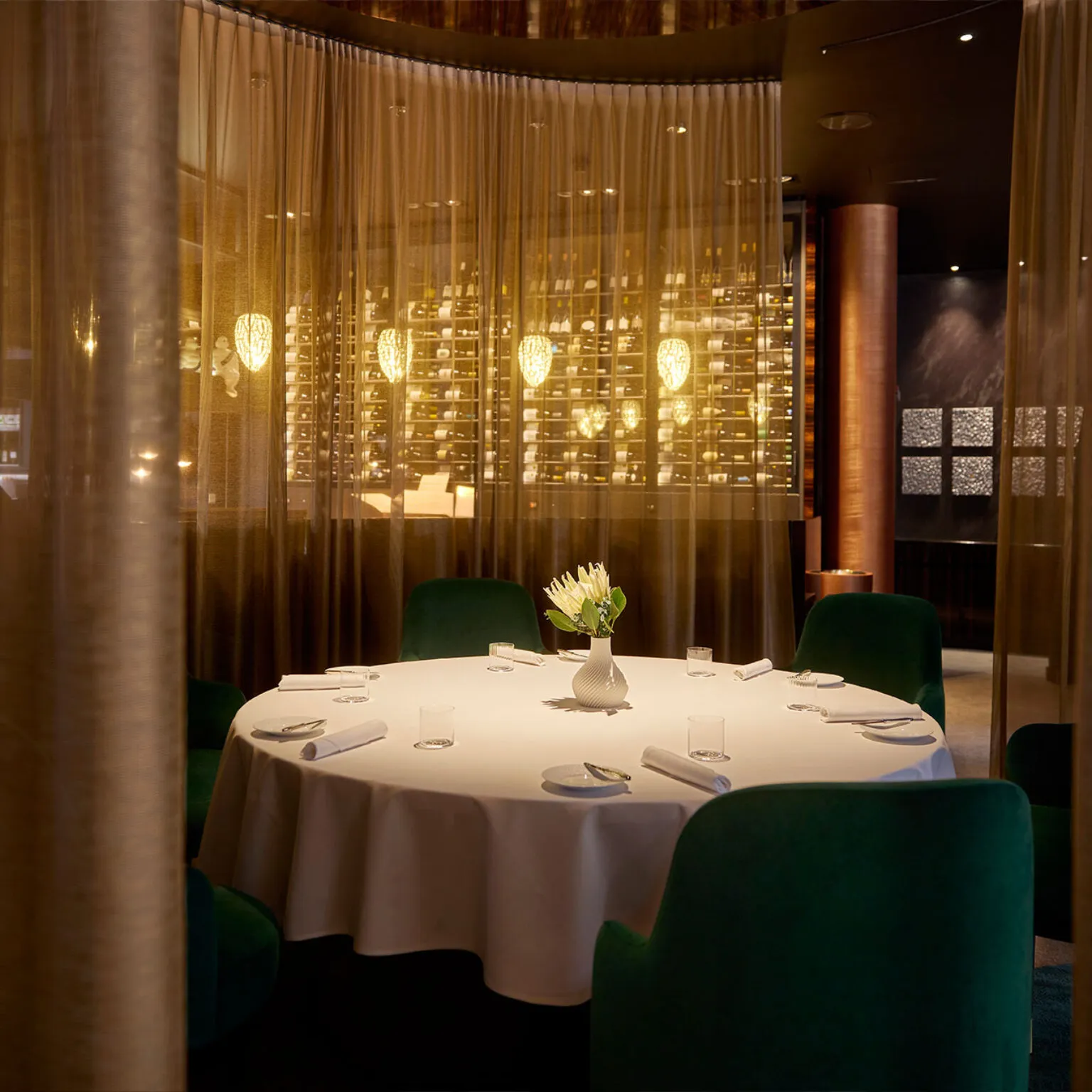 Innenansicht des Restaurants Ammolite – House of Light. Ein weiß gedeckter Tisch mit einer Blumenvase im Vordergrund. Im Hintergrund feine, durchsichtige Gardinen. Man sieht die edle Beleuchtung durchschimmern.