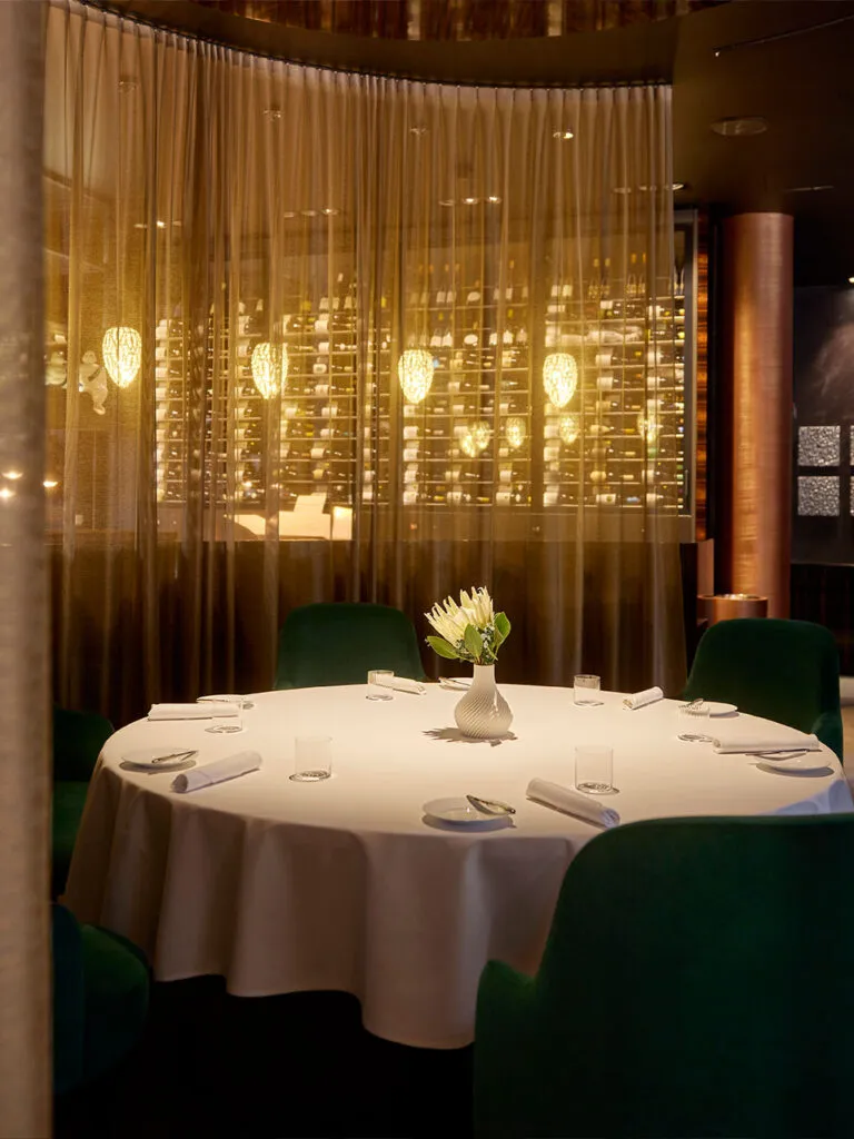 Innenansicht des Restaurants Ammolite – House of Light. Ein weiß gedeckter Tisch mit einer Blumenvase im Vordergrund. Im Hintergrund feine, durchsichtige Gardinen. Man sieht die edle Beleuchtung durchschimmern.