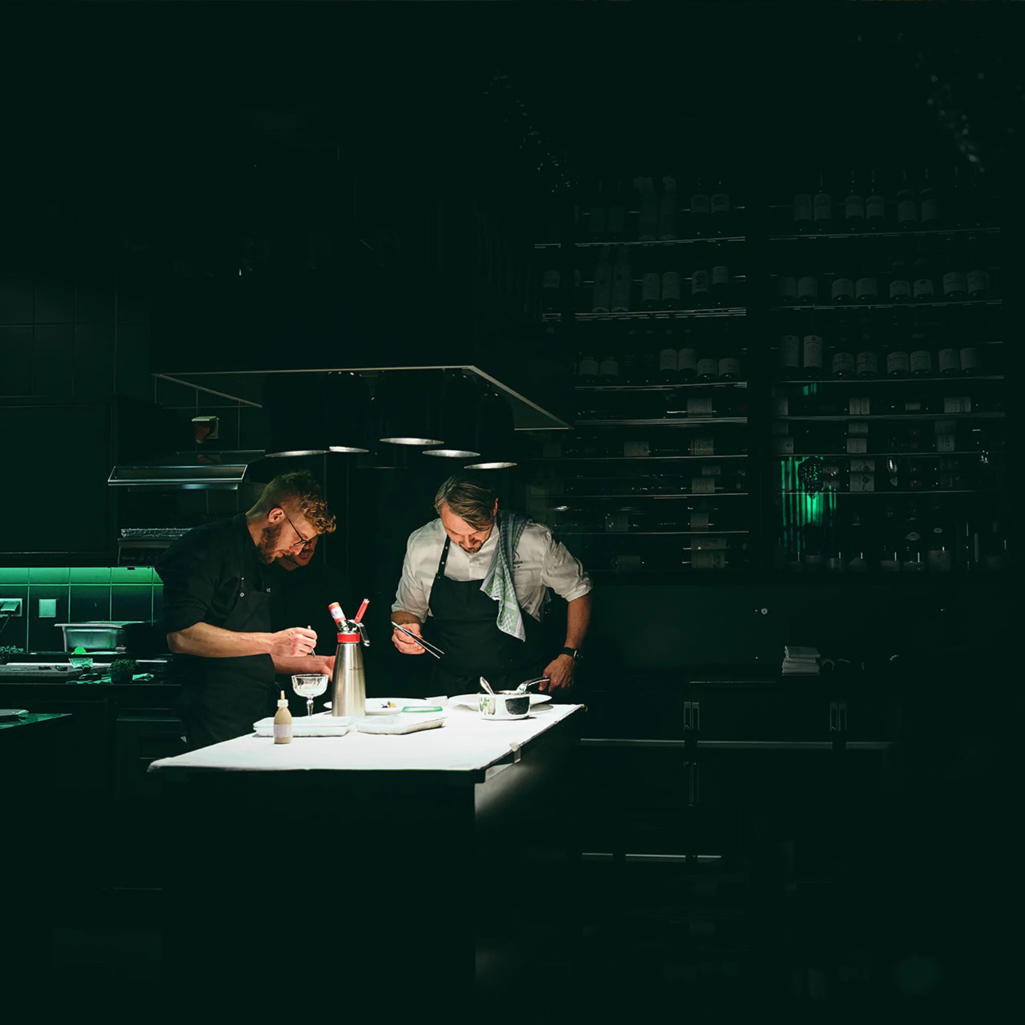 Peter Hagen-Wiest richtet mit seinem Team in der Küche des Ammolite ein Menü auf einer beleuchteten weißen Arbeitsfläche. Der Raum ist, bis auf den Tisch, dunkel.