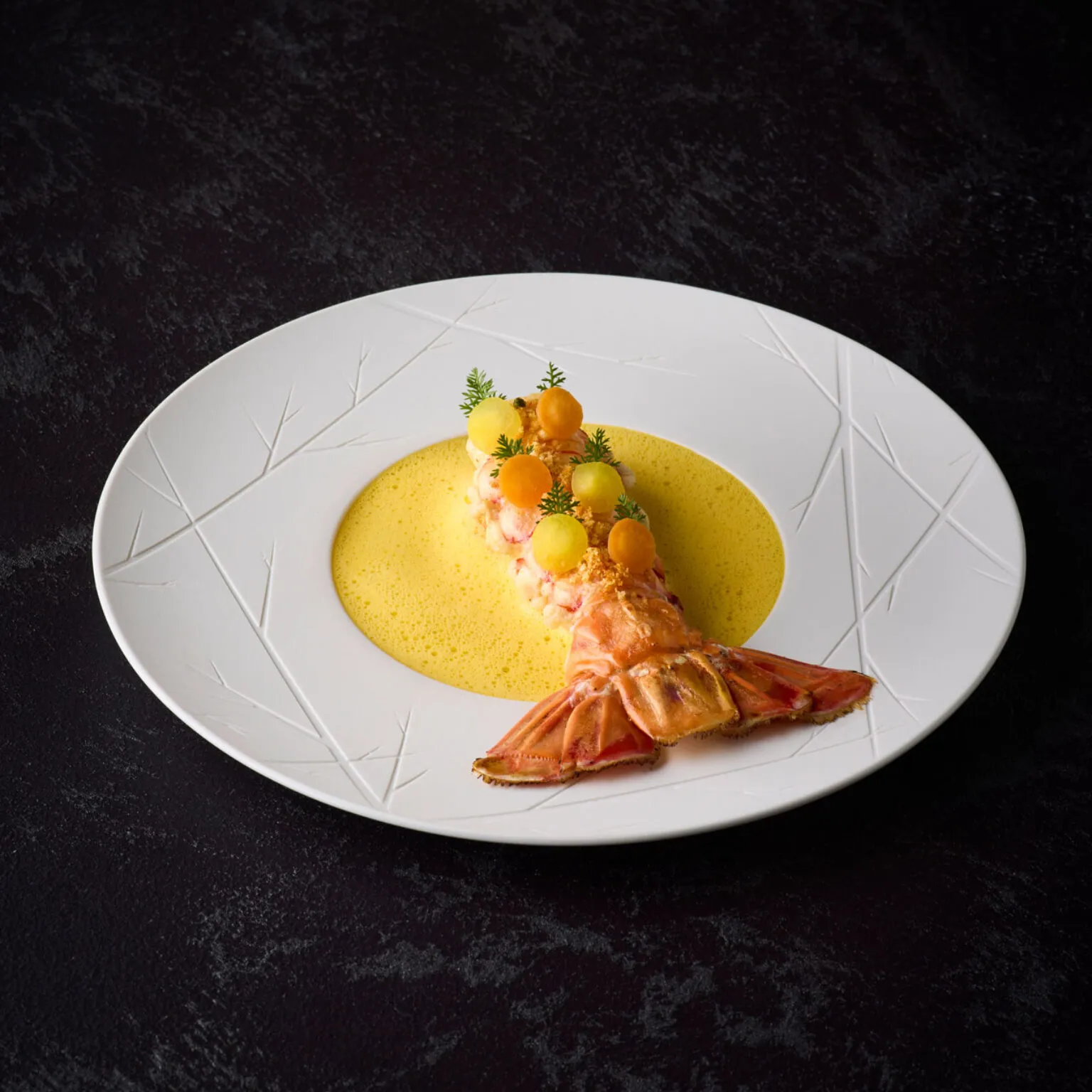 Ein Kaisergranatschwanz serviert mit gelben und orangen Karottenscheiben auf einem weißen Teller. Darunter sieht man eine gelbe Schaumsoße.