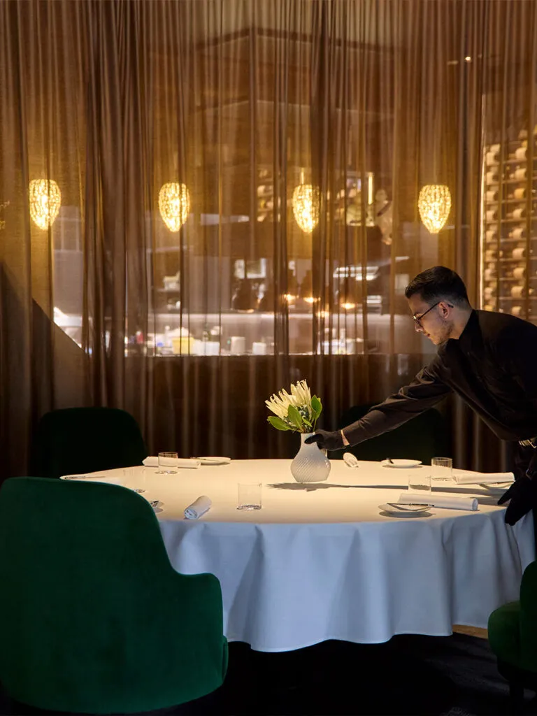 Ein Teammitglied des Ammolite – House of Light deckt einen Tisch im Restaurant ein. Er stellt gerade eine Blumenvase auf den Tisch.