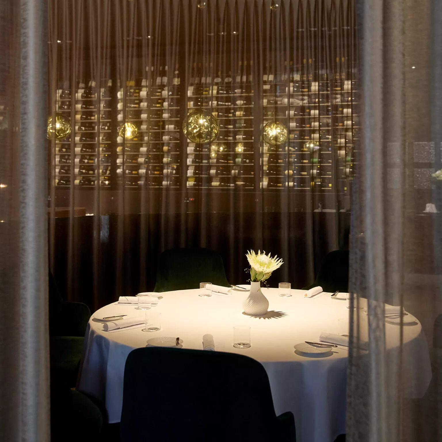 Vue intérieure du restaurant Ammolite – House of Light. Une table blanche dressée avec un vase fleuri au premier plan. À l’arrière-plan, des rideaux fins et transparents. On devine l’éclairage d’ambiance raffiné derrière.